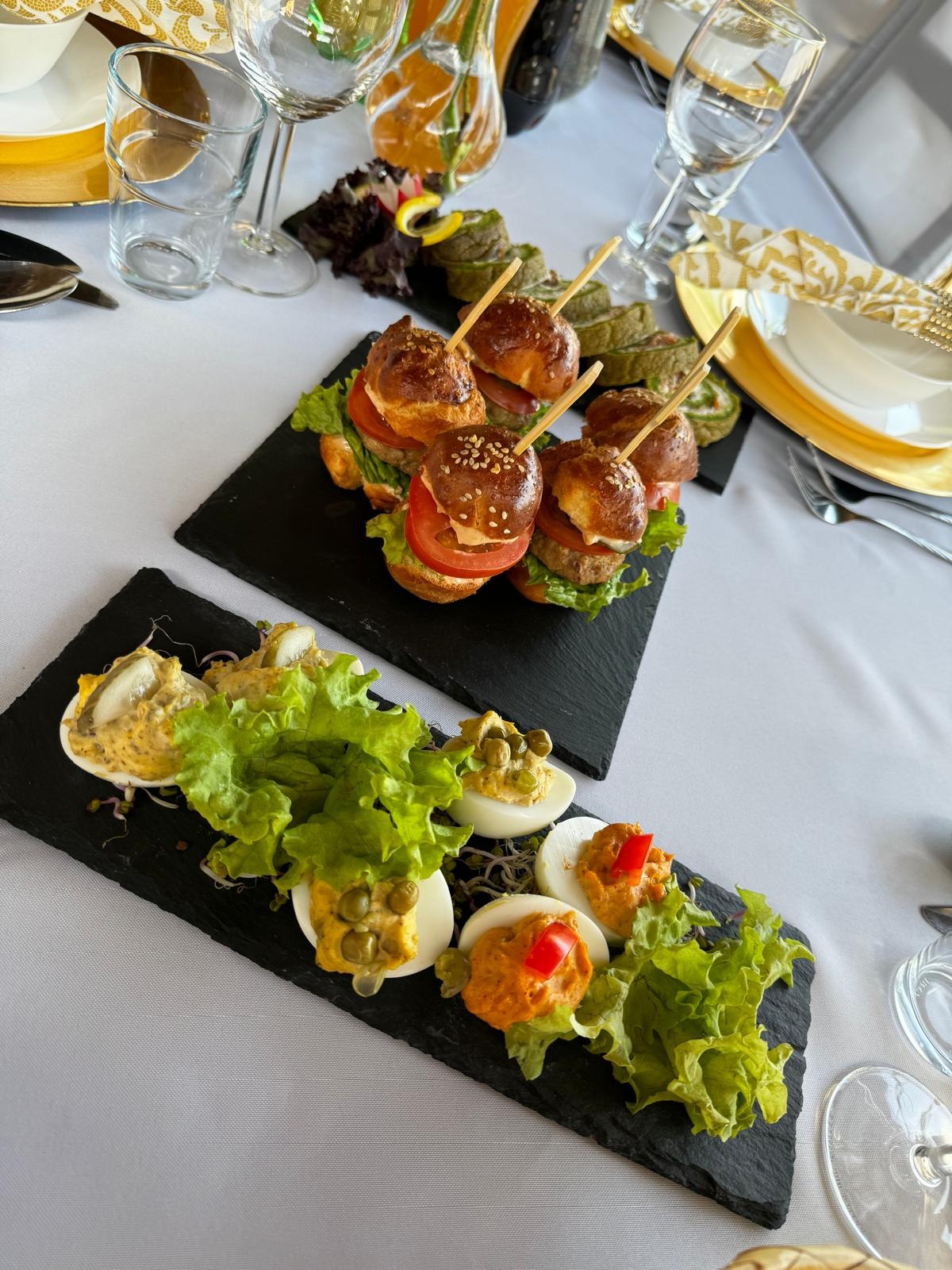 Elegancko podane przekąski na czarnych łupkach: mini burgery z wykałaczkami, faszerowane jajka z dekoracją z sałaty, zielone roladki. Nakryty stół z białym obrusem i złotymi akcentami.