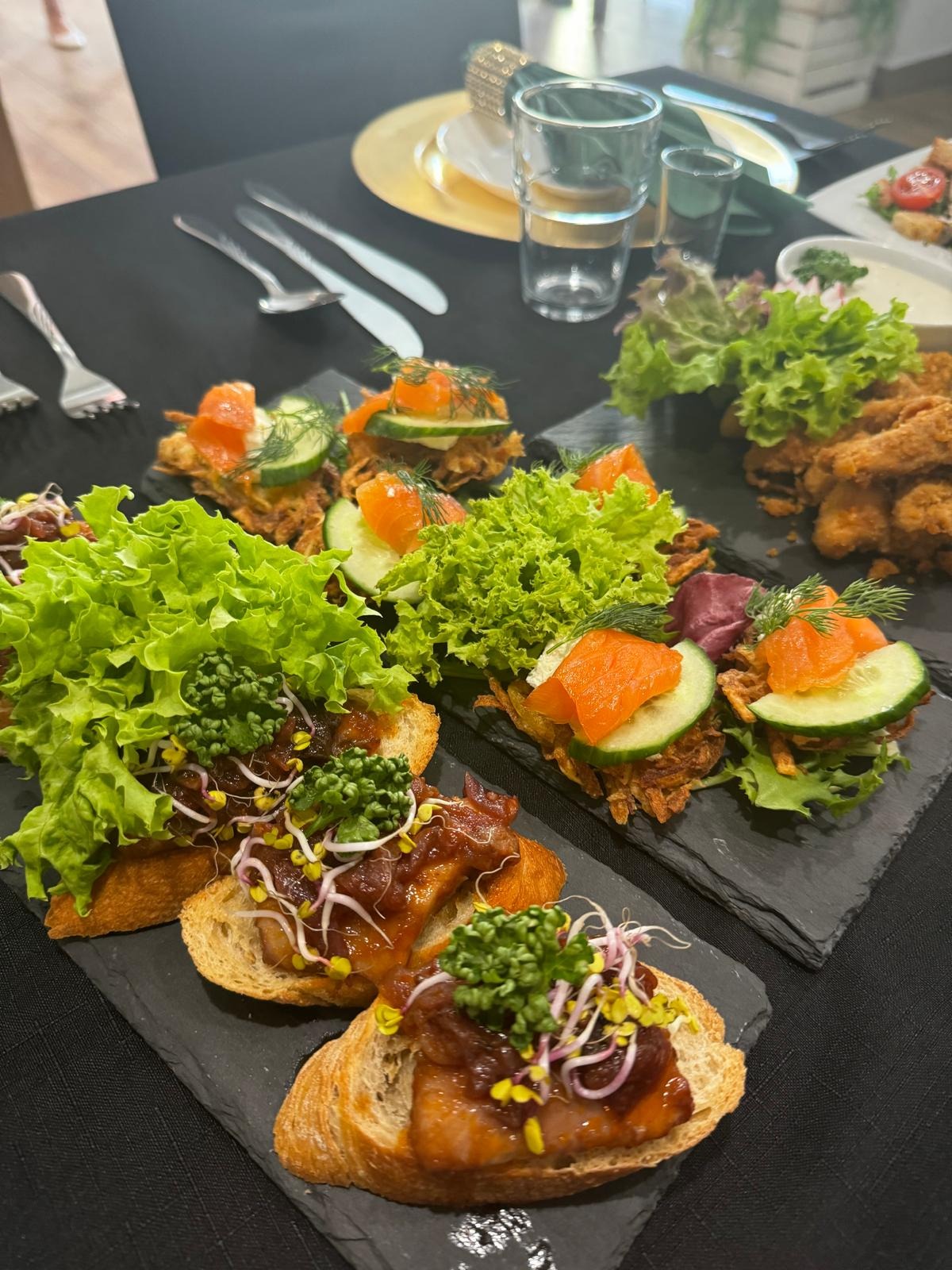Elegancko podane kanapeczki na ciemnych kamiennych deskach: jedne z łososiem i ogórkiem, inne z mięsem, sałatą i kiełkami, na czarnym obrusie, z widelcami i szklankami w tle.