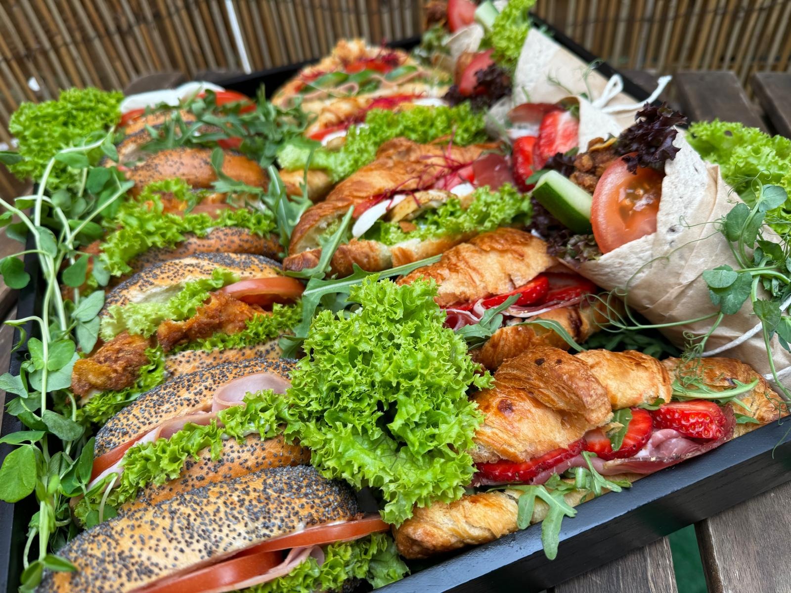 Taca z różnorodnymi kanapkami i wrapami, w tym bagietki z makiem, croissanty z szynką i truskawkami, oraz zawijasy z warzywami, przygotowana przez firmę z Radomia.
