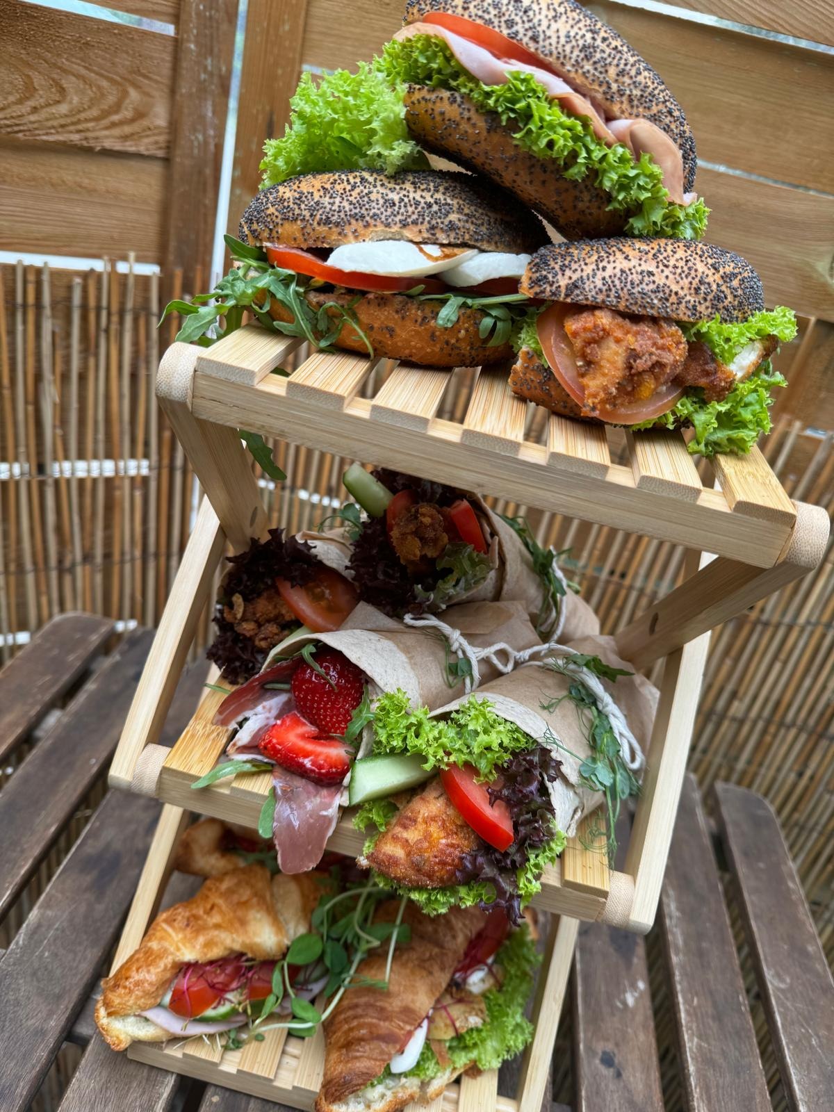 Trzypoziomowy drewniany stojak prezentuje różnorodne przekąski: kanapki na bułkach posypanych makiem, tortille zawinięte w papier oraz croissanty z dodatkami, wszystko udekorowane świeżymi...