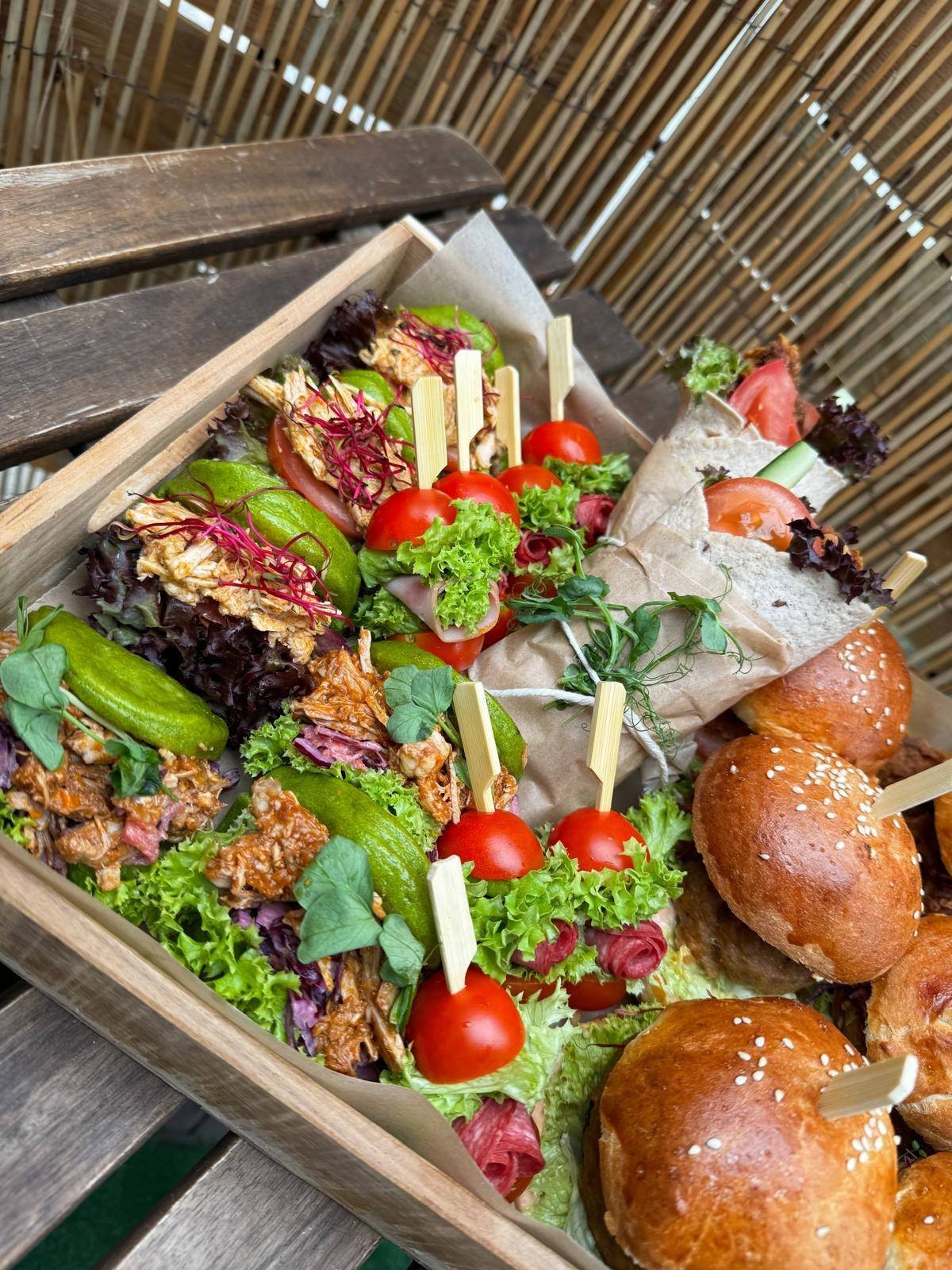 Drewniana skrzynka z różnorodnymi przekąskami: mini burgery z sezamem, roladki w papierze, koreczki z pomidorkami cherry i sałatą, zielone papryczki faszerowane, całość na podkładzie z sałaty.