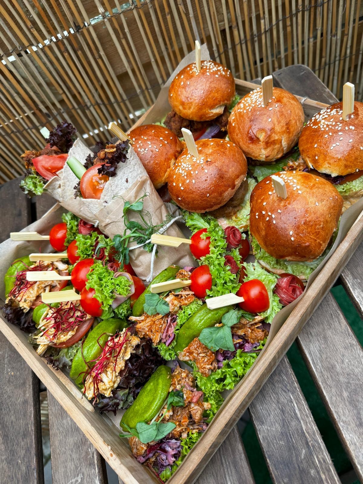 Drewniana taca z mini burgerami z sezamem, wrapami w papierze kraft i przekąskami na szpadkach, udekorowanymi pomidorkami cherry i zieleniną, na tle bambusowej maty.