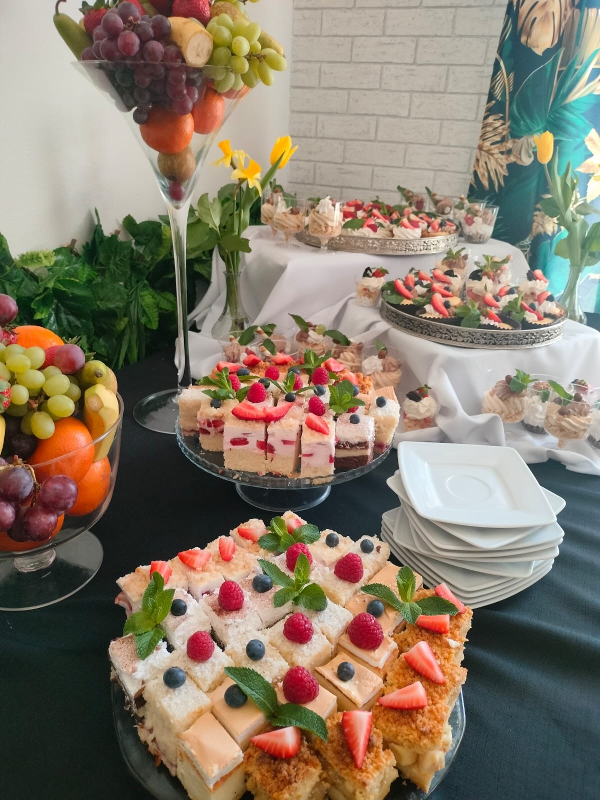 Stół cateringowy z różnorodnymi deserami: mini torciki z truskawkami i borówkami, desery w szklankach, owoce w wazonach, dekorowany kwiatami i zielenią.