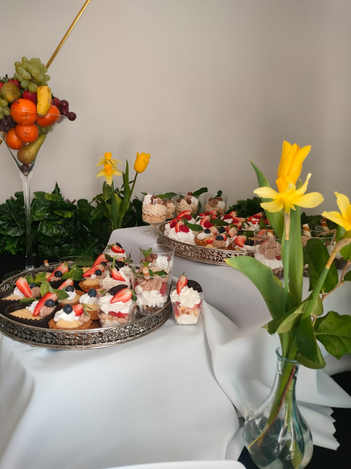 Stół z deserami: tartaletki z owocami, mini desery w kubeczkach, taca z ciastami, dekoracja z owoców w kielichu i żółte tulipany w wazonie.
