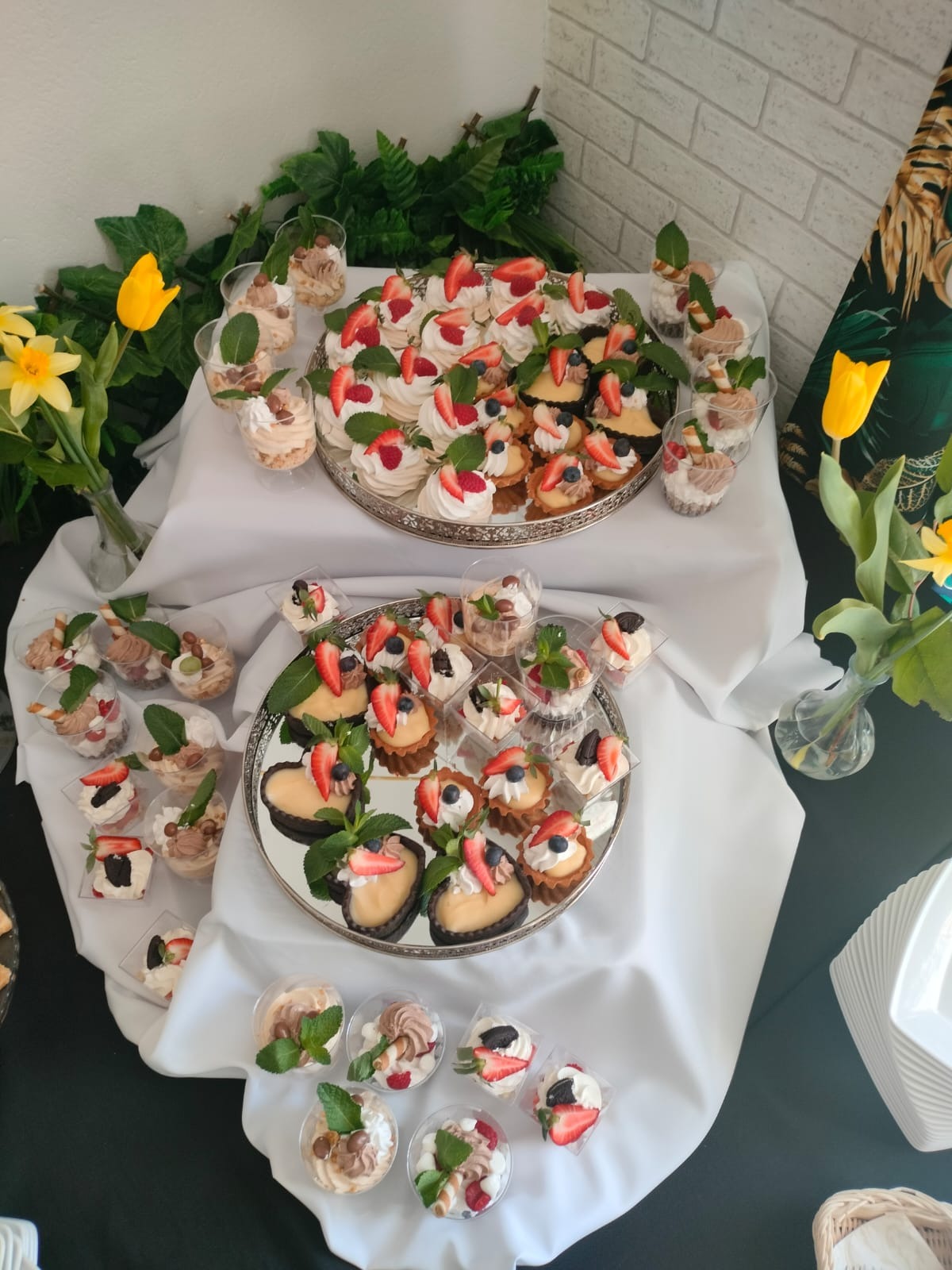 Elegancko zaaranżowany stół z deserami: mini bezy z owocami, tartaletki z kremem i owocami oraz desery w przezroczystych kubeczkach, udekorowane miętą, na srebrnych tacach, z kwiatami w tle.