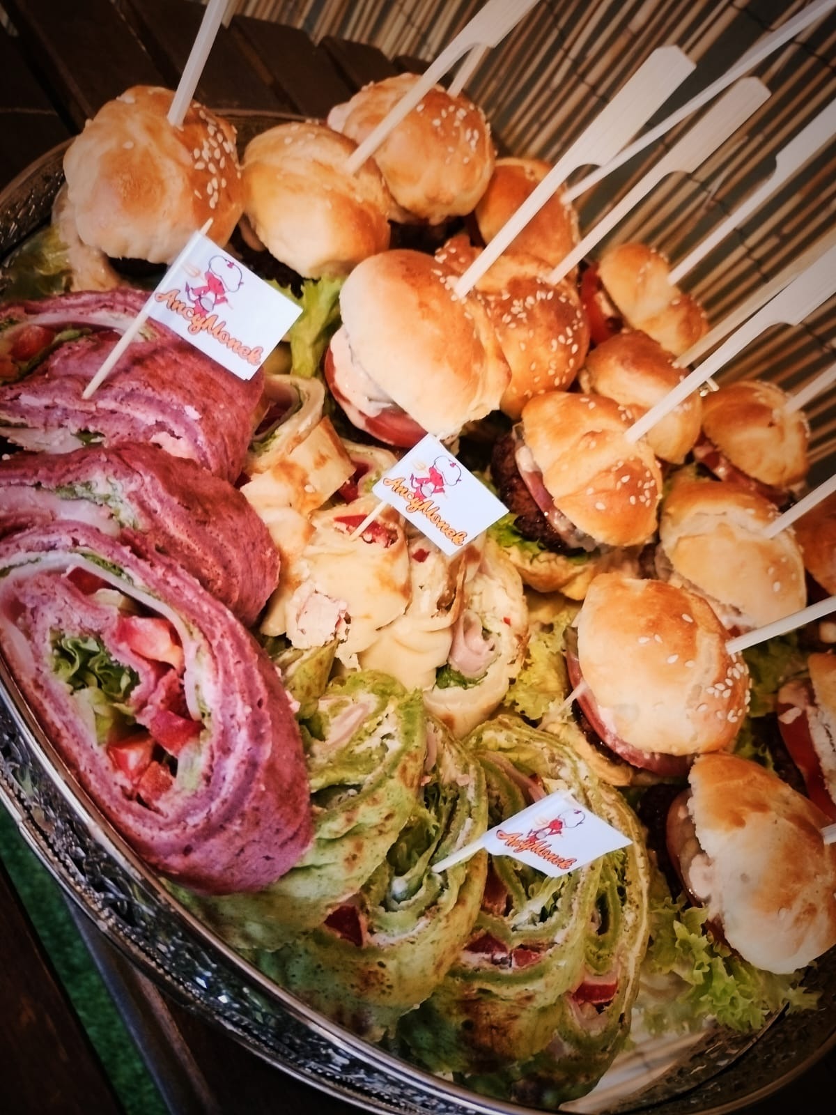Srebrna taca z kolorowymi roladkami z nadzieniem, mini burgerami i przekąskami na przyjęcie, ozdobiona flagami z logo firmy.