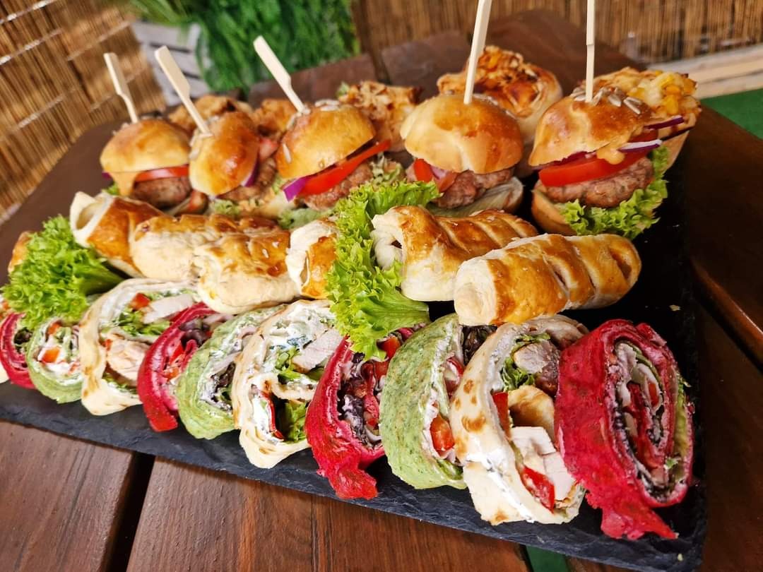 Elegancko ułożone na czarnym talerzu mini burgery z wykałaczkami, roladki z kolorowych tortilli z różnymi nadzieniami oraz przekąski z ciasta francuskiego.