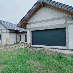 Fhu De-ko - Garaż z ciemnozieloną bramą segmentową, budynek w stanie surowym, białe bloczki, czarna dachówka, drewniane elementy wykończeniowe dachu, teren trawiasty.