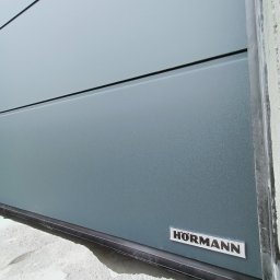 Fhu De-ko - Fragment szarej bramy garażowej z widocznym logo producenta, zamontowanej w surowym stanie budynku.