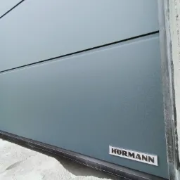 Fragment szarej bramy garażowej z widocznym logo producenta, zamontowanej w surowym stanie budynku.