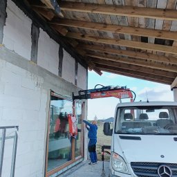 Fhu De-ko - Montaż okna za pomocą przyssawki i dźwigu zamontowanego na samochodzie dostawczym, widoczny pracownik w niebieskim ubraniu oraz surowy stan budynku z widoczną konstrukcją dachu.