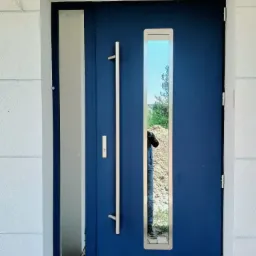 Nowoczesne, niebieskie drzwi wejściowe z długim, pionowym uchwytem i prostokątnym okienkiem, w którym odbija się sylwetka osoby i fragment krajobrazu, osadzone w białej, teksturowanej ścianie.