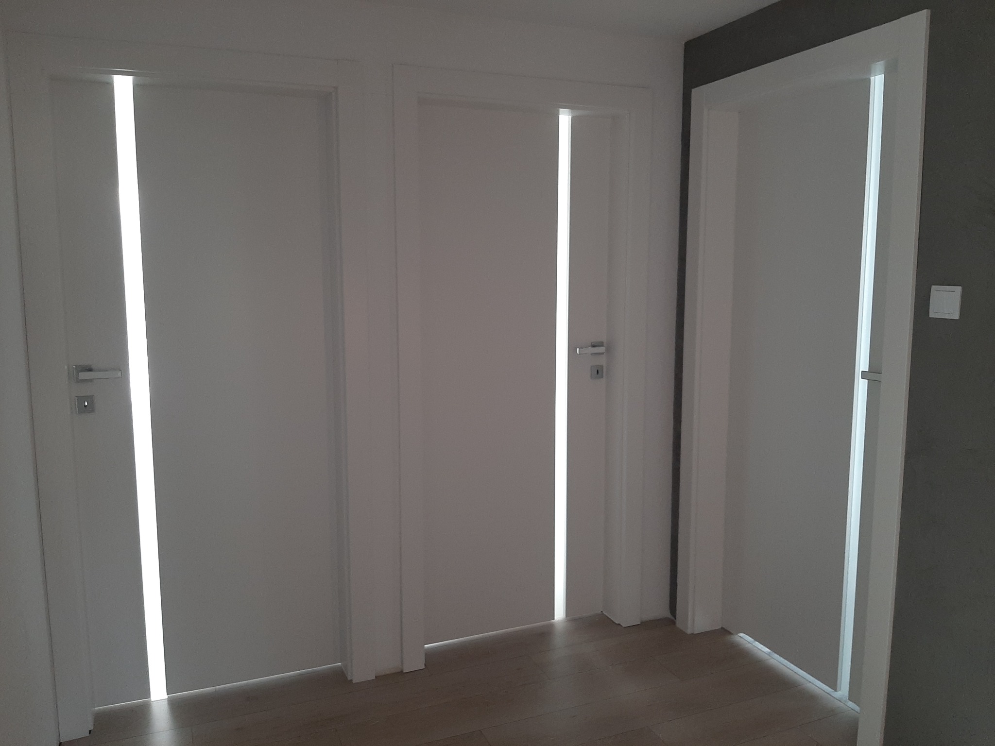 Trzy białe, minimalistyczne drzwi wewnętrzne z podłużnymi przeszkleniami, widoczne klamki i fragment podłogi.