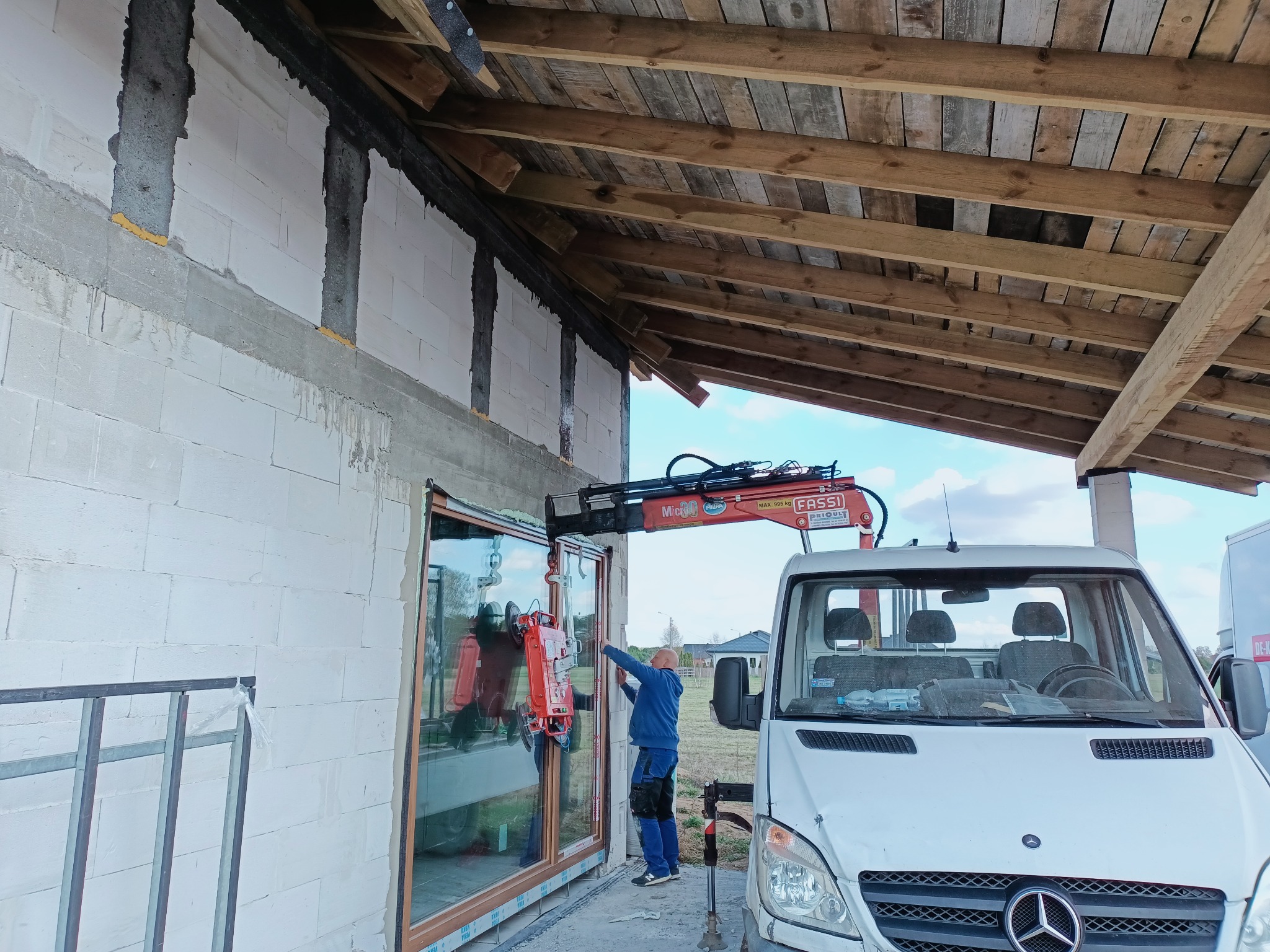 Montaż okna za pomocą przyssawki i dźwigu zamontowanego na samochodzie dostawczym, widoczny pracownik w niebieskim ubraniu oraz surowy stan budynku z widoczną konstrukcją dachu.