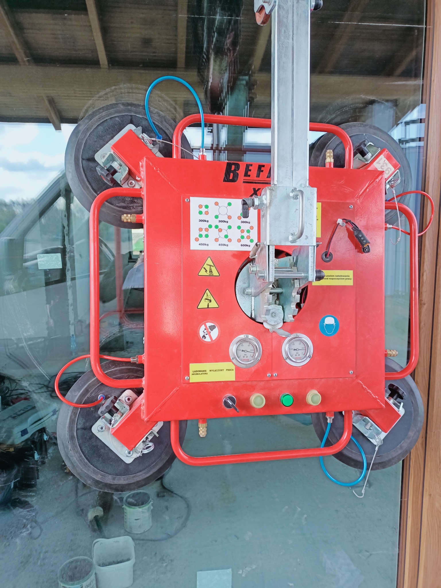 Czerwony manipulator próżniowy z przyssawkami mocowany do szyby okiennej podczas montażu, widoczne manometry i schemat obciążenia urządzenia.