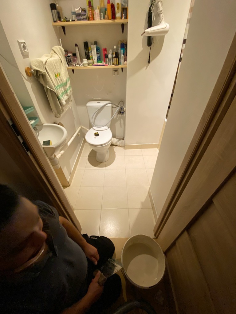 Widok z góry na małą łazienkę z beżowymi płytkami podłogowymi, z mężczyzną siedzącym na podłodze obok wiadra z szarą cieczą, prawdopodobnie przygotowującego podłoże do dalszych prac wykończeniowych.