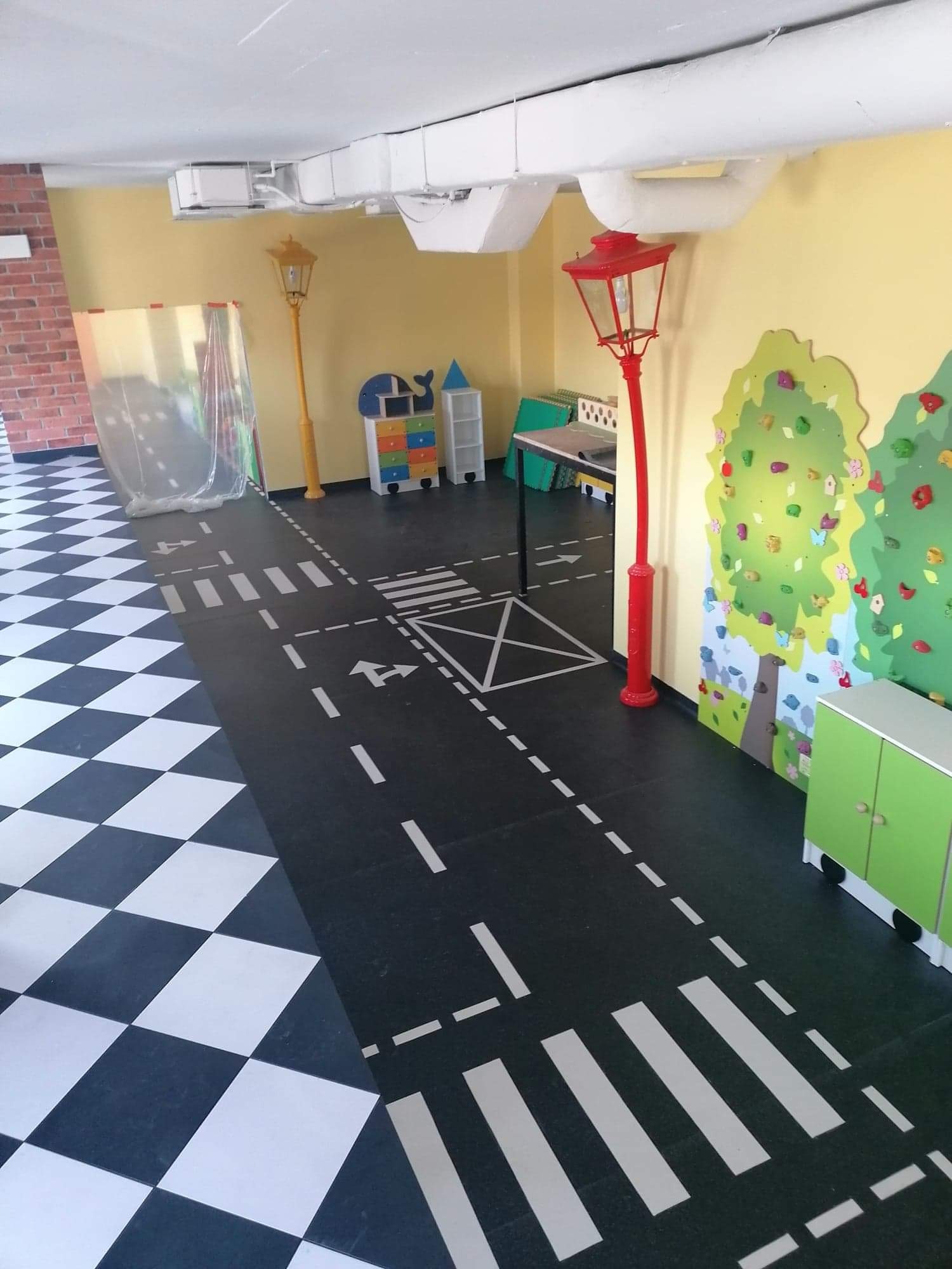 Pokój dziecięcy z podłogą imitującą ulicę, z czarno-białą szachownicą przechodzącą w asfalt z namalowanymi pasami, lampami ulicznymi i ścianą wspinaczkową w kształcie drzewa.