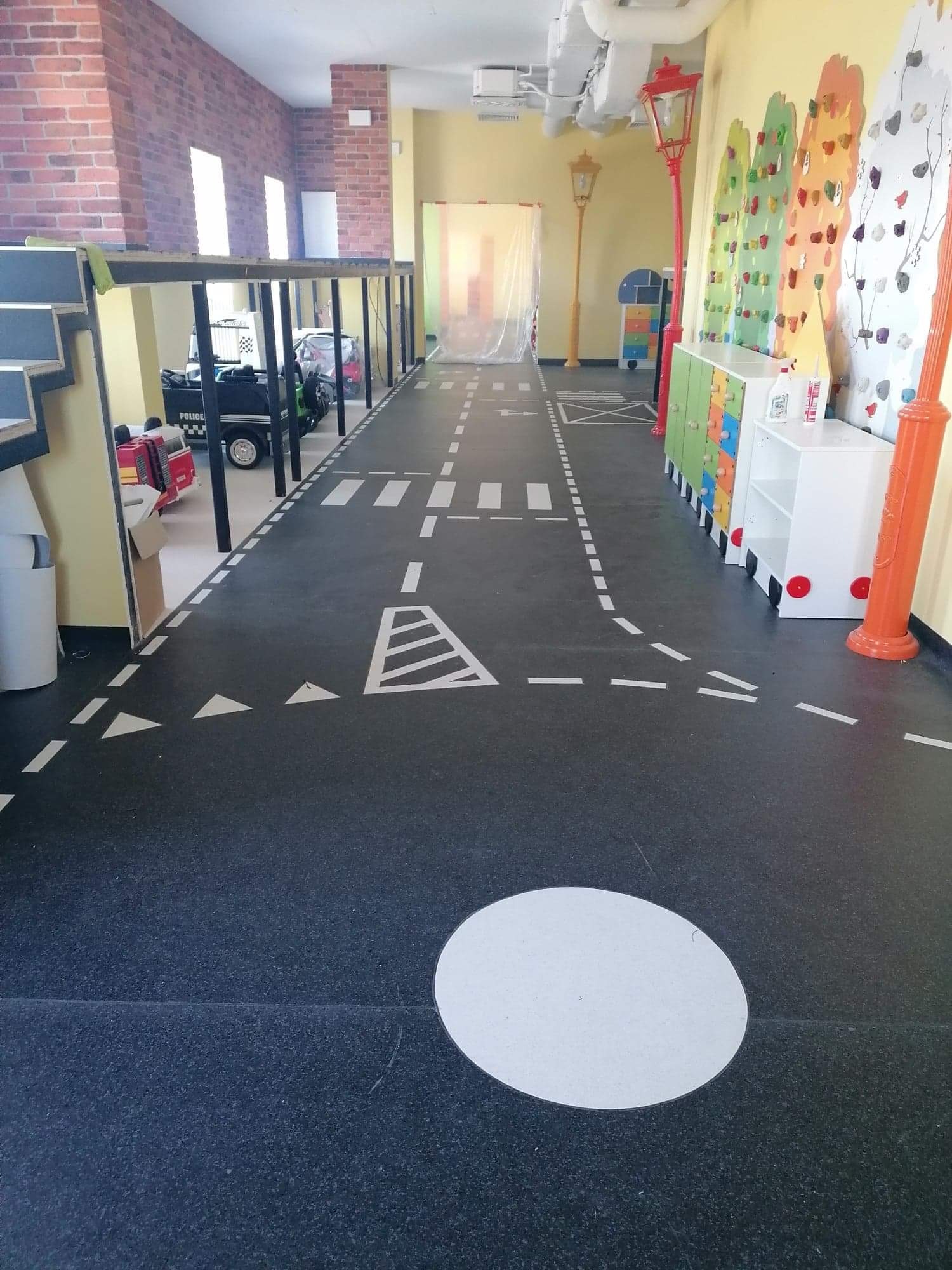 Podłoga w pokoju zabaw z czarnej wykładziny imitującej asfalt, pomalowana w białe pasy i znaki drogowe, z elementami dekoracyjnymi jak lampy uliczne i ściana wspinaczkowa w tle.