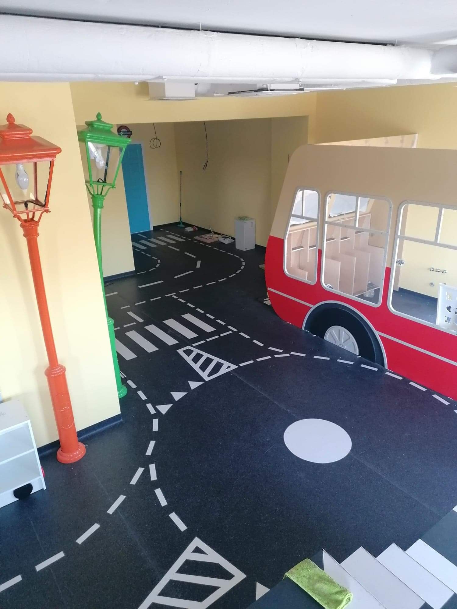 Pokój zabaw z podłogą imitującą ulicę, z namalowanymi przejściami dla pieszych i drogami, czerwony autobus-zabawka i dwa stylizowane lampiony: pomarańczowy i zielony.