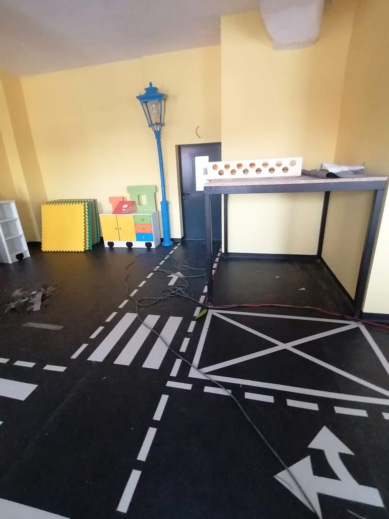 Pokój dziecięcy z podłogą imitującą ulicę z pasami i strzałkami, żółte ściany, zabawki i lampa uliczna; widoczne kable na podłodze.