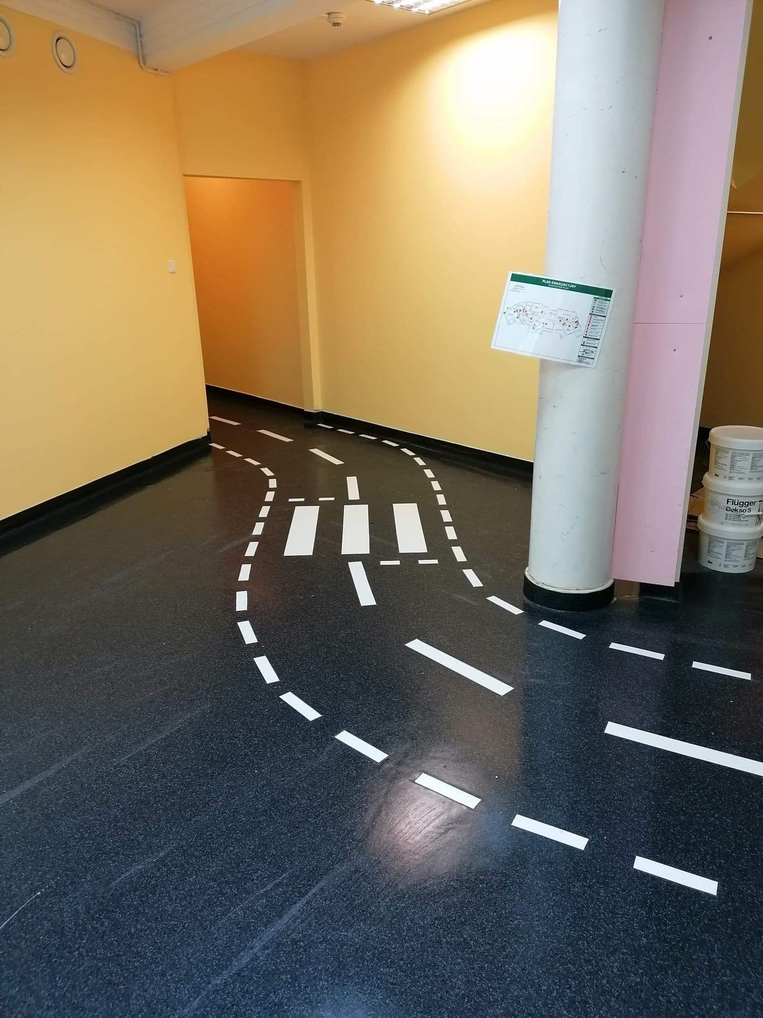 Korytarz z czarną podłogą, na której wymalowano białe linie tworzące imitację drogi i przejścia dla pieszych; na ścianie wisi plan ewakuacyjny, a w rogu stoją trzy pojemniki z farbą.