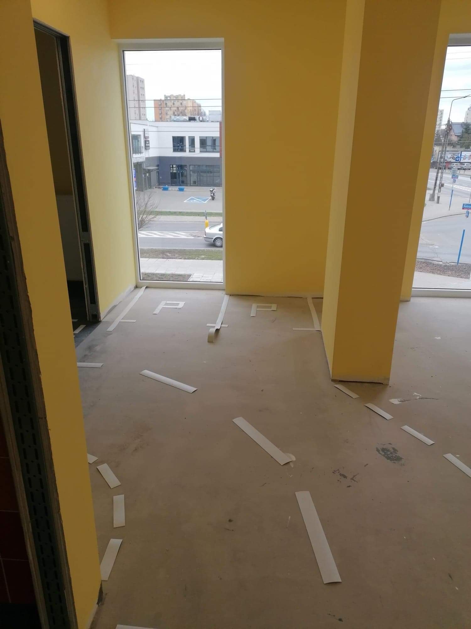 Przygotowanie do montażu wykładziny w biurze: fragment betonowej podłogi z taśmami wyznaczającymi układ, żółte ściany i widok na ulicę przez okno.
