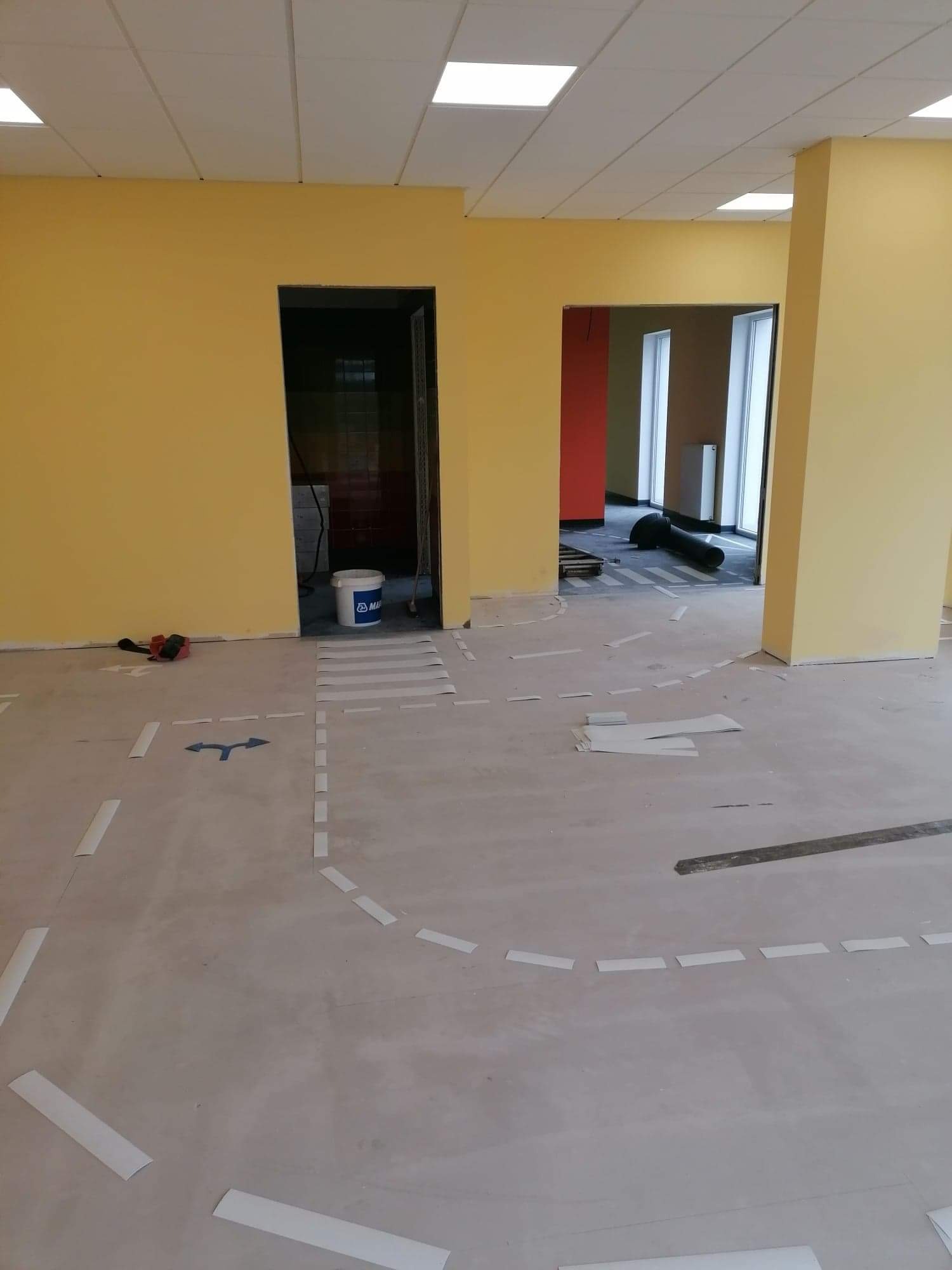 Przygotowanie podłoża w biurze z żółtymi ścianami, białe paski wyznaczają obszar, wiadro z klejem i szlifierka przy ścianie, w tle widoczne rolki wykładziny