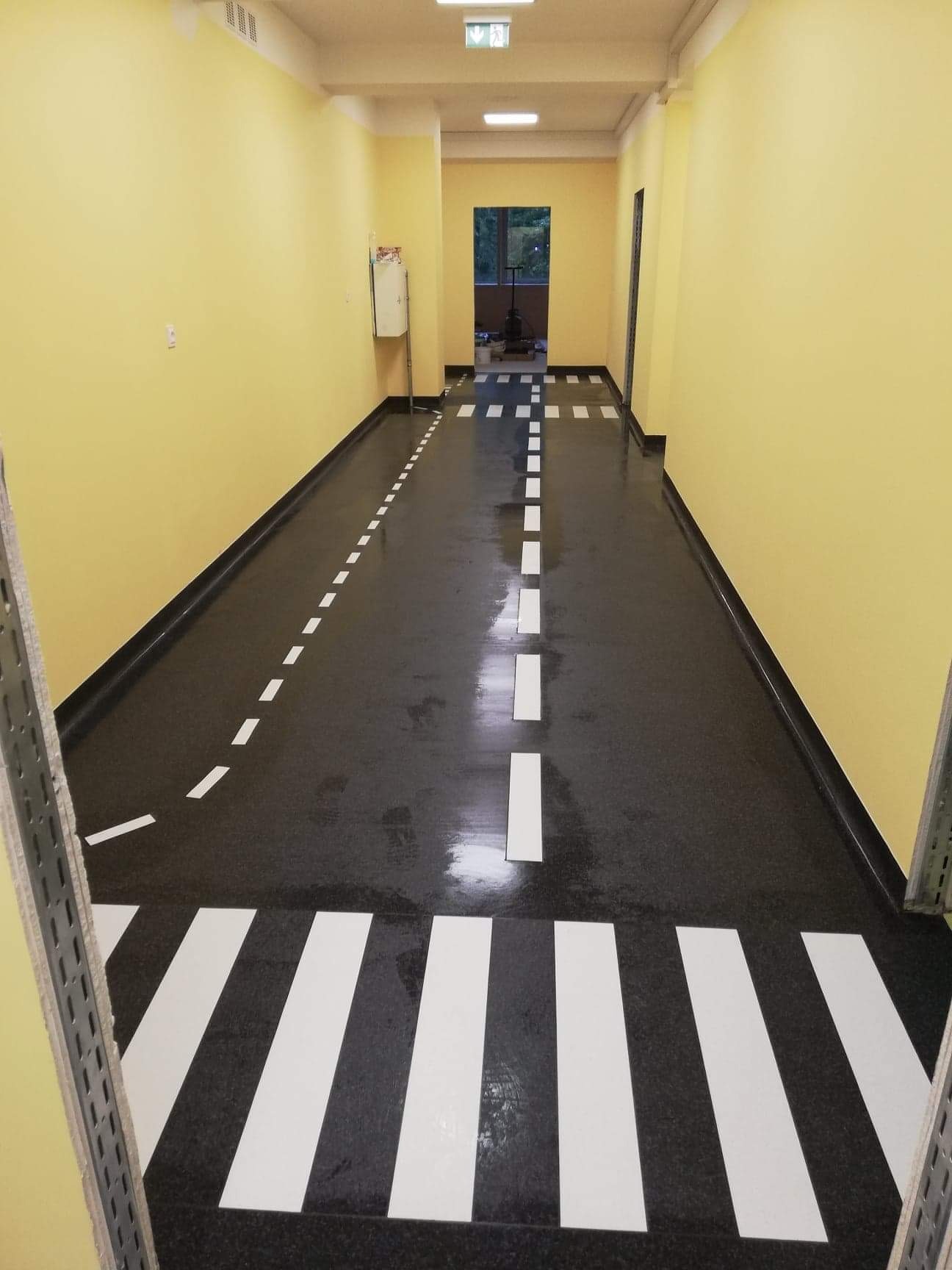 Korytarz z czarną podłogą imitującą jezdnię z białymi pasami i liniami, żółte ściany, widok na dalszą część korytarza z oknem.