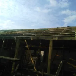 Konstrukcja drewniana dachu w trakcie budowy, widoczne łaty i krokwie, na tle błękitnego nieba z chmurami.