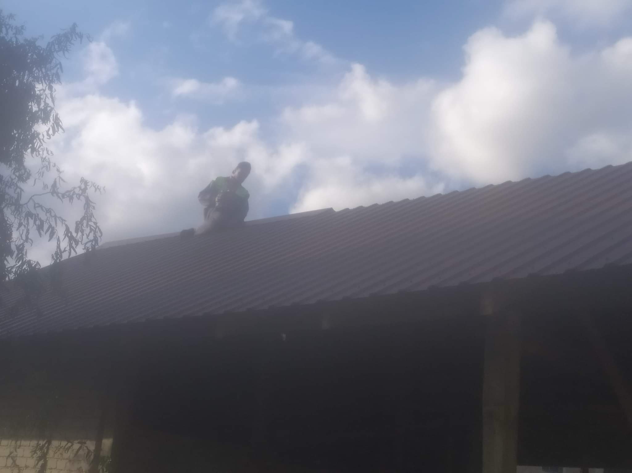 Pracownik w zielonej kurtce montuje ciemną blachę trapezową na dachu budynku gospodarczego, widoczne jasne chmury na niebie.