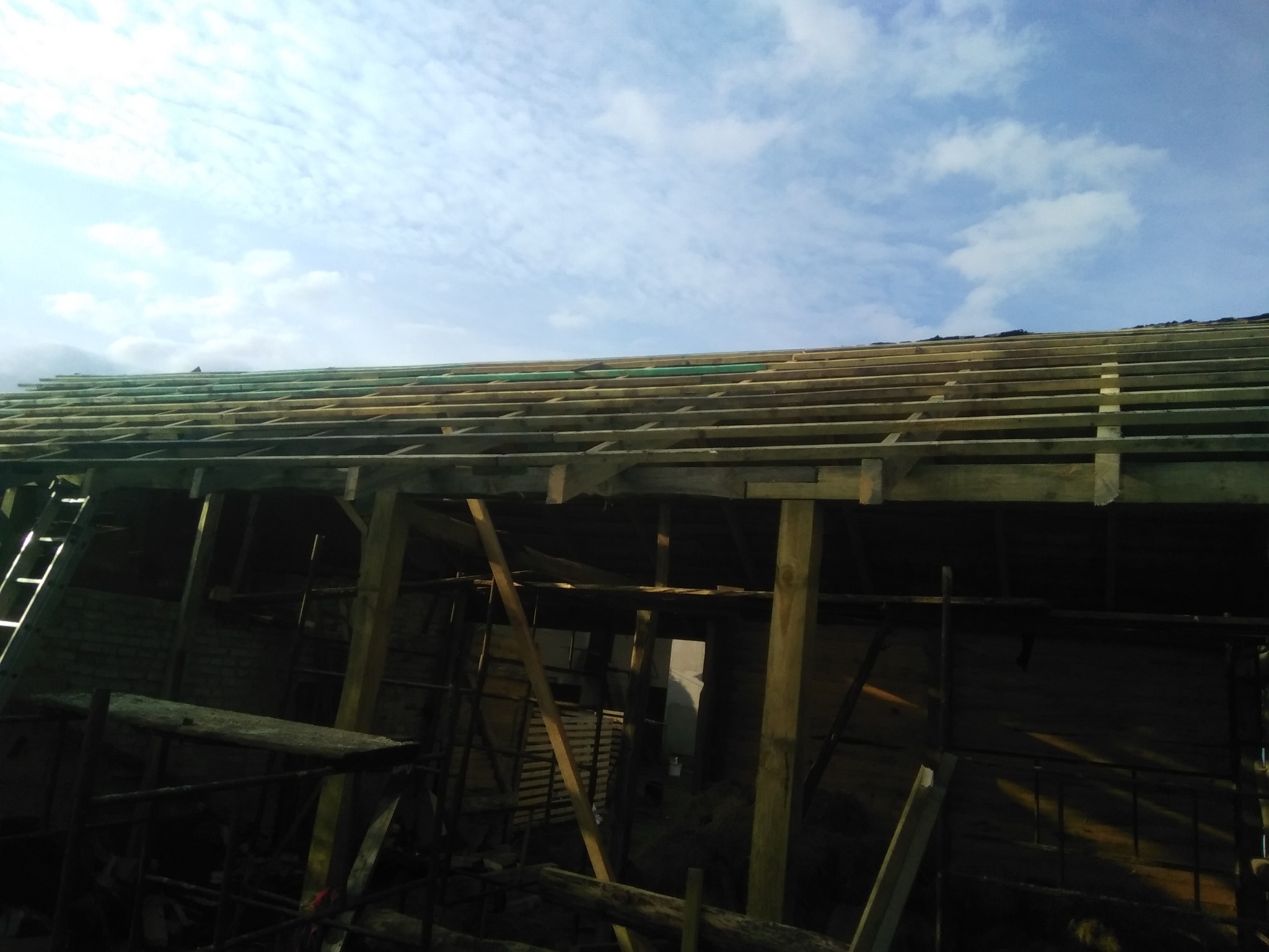 Konstrukcja drewniana dachu w trakcie budowy, widoczne łaty i krokwie, na tle błękitnego nieba z chmurami.