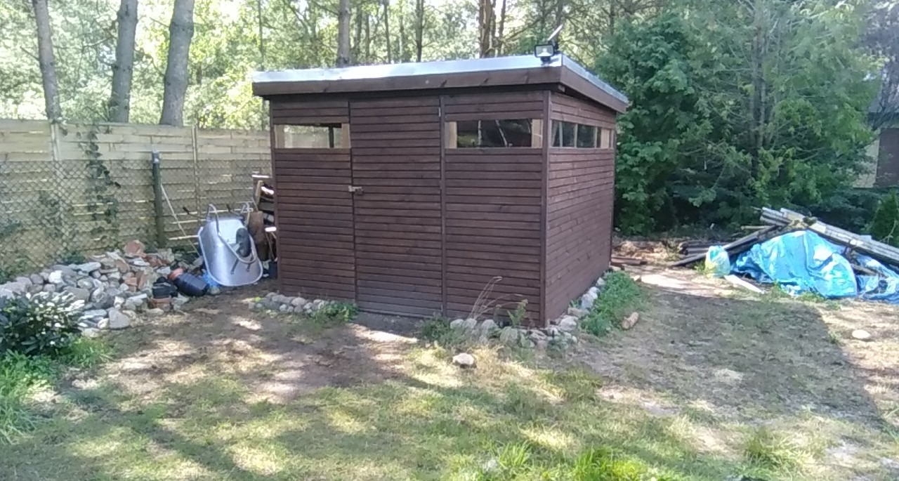 Brązowy drewniany domek narzędziowy z oknami i metalowym dachem, oświetlony lampą solarną, otoczony kamieniami i gruzem w ogrodzie.