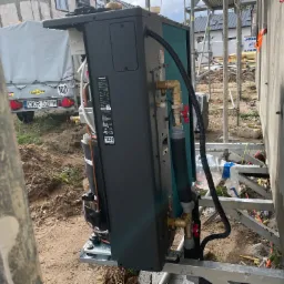 Czarna jednostka zewnętrzna pompy ciepła Panasonic, umieszczona na metalowej platformie przy ścianie budynku w trakcie montażu, widoczne rury i zawory, tło budowlane z przyczepą i rusztowaniami.