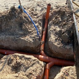 SONO Sp.z o.o. - Pomarańczowe rury instalacyjne PVC z kolankami i trójnikami, ułożone w piaszczystym wykopie. Widoczny pionowy odcinek i niebieski przewód.