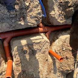 SONO Sp.z o.o. - Instalacja pomarańczowych rur kanalizacyjnych z rozgałęzieniami w ziemnym wykopie, obok niebieski przewód i fragment stopy pracownika. Widoczna czarna ściana fundamentowa z czerwoną linką.