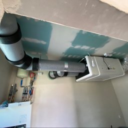 SONO Sp.z o.o. - Centrala wentylacyjna z rekuperacją, podwieszona do sufitu z płyt GK, połączona z szarymi kanałami. Obok rozdzielacz hydrauliczny z naczyniem wzbiorczym, w pomieszczeniu w trakcie wykańczania.