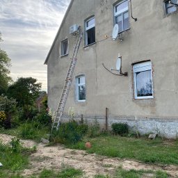 SONO Sp.z o.o. - Zewnętrzna jednostka klimatyzacji zamontowana na szarej, popękanej ścianie starego budynku, z metalową drabiną opartą o elewację, w tle zachmurzone niebo.