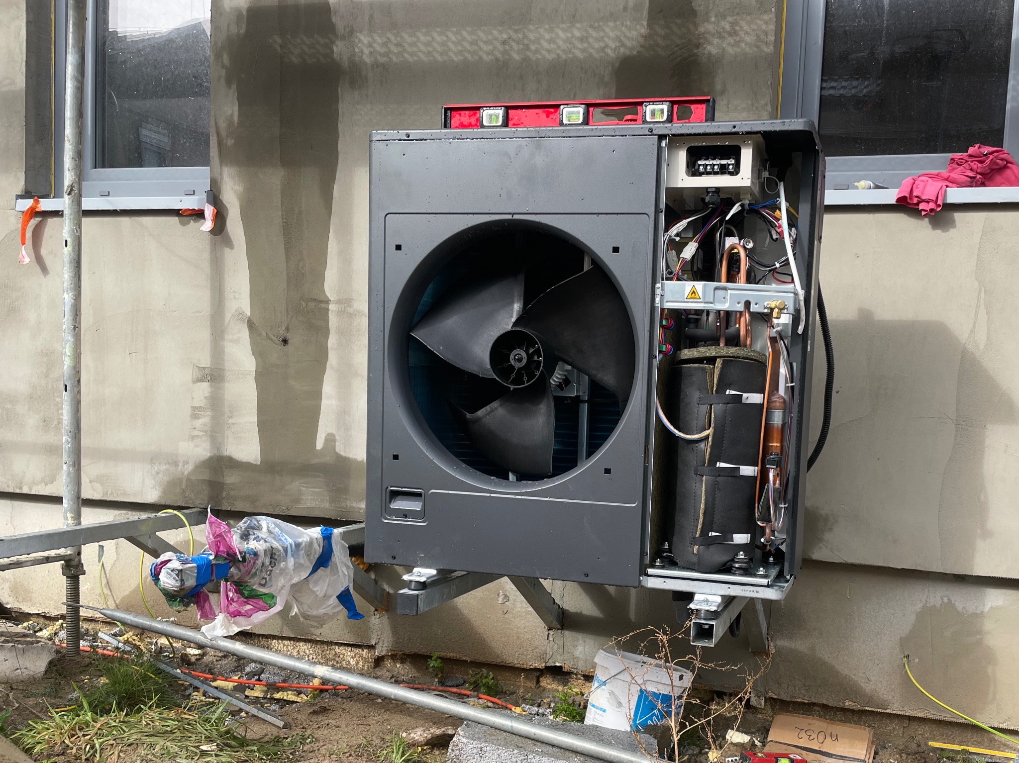 Szara jednostka zewnętrzna z otwartą obudową, widoczne elementy instalacji, poziomica na górze, montaż na rusztowaniu przy ścianie budynku w trakcie prac wykończeniowych.