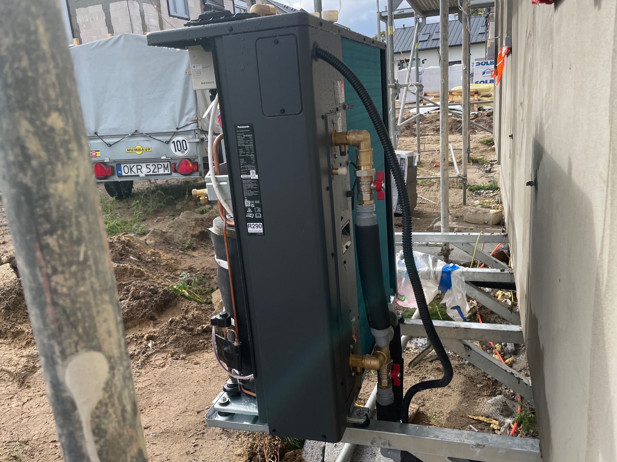 Czarna jednostka zewnętrzna pompy ciepła Panasonic, umieszczona na metalowej platformie przy ścianie budynku w trakcie montażu, widoczne rury i zawory, tło budowlane z przyczepą i rusztowaniami.
