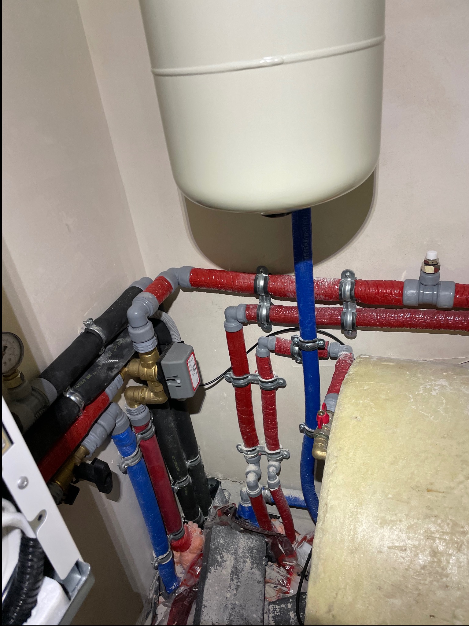 Węzeł instalacji grzewczej z widocznym zbiornikiem wyrównawczym, rurami w izolacji czerwonej oraz niebieskiej, zaworami i osprzętem w pomieszczeniu technicznym.