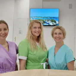 Trzy uśmiechnięte kobiety w kolorowych uniformach medycznych stojące za jasnym biurkiem w gabinecie, z telewizorem wyświetlającym krajobraz i słonecznikiem w wazonie w tle.
