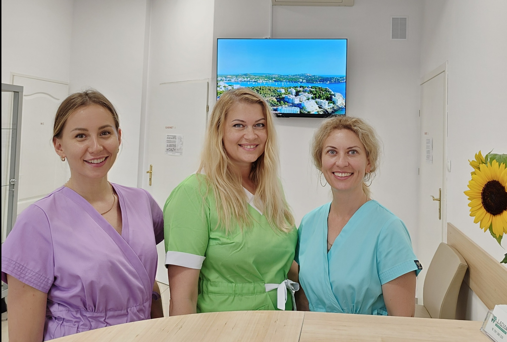 Trzy uśmiechnięte kobiety w kolorowych uniformach medycznych stojące za jasnym biurkiem w gabinecie, z telewizorem wyświetlającym krajobraz i słonecznikiem w wazonie w tle.