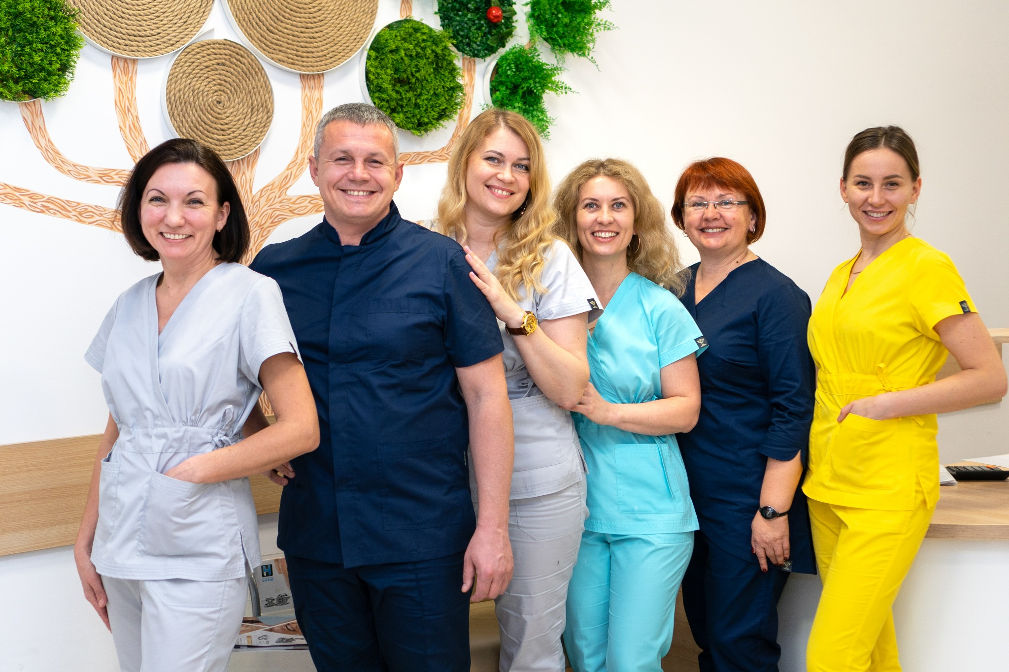 Zespół lekarzy w kolorowych uniformach pozuje w jasnym gabinecie z dekoracją ścienną imitującą drzewo.