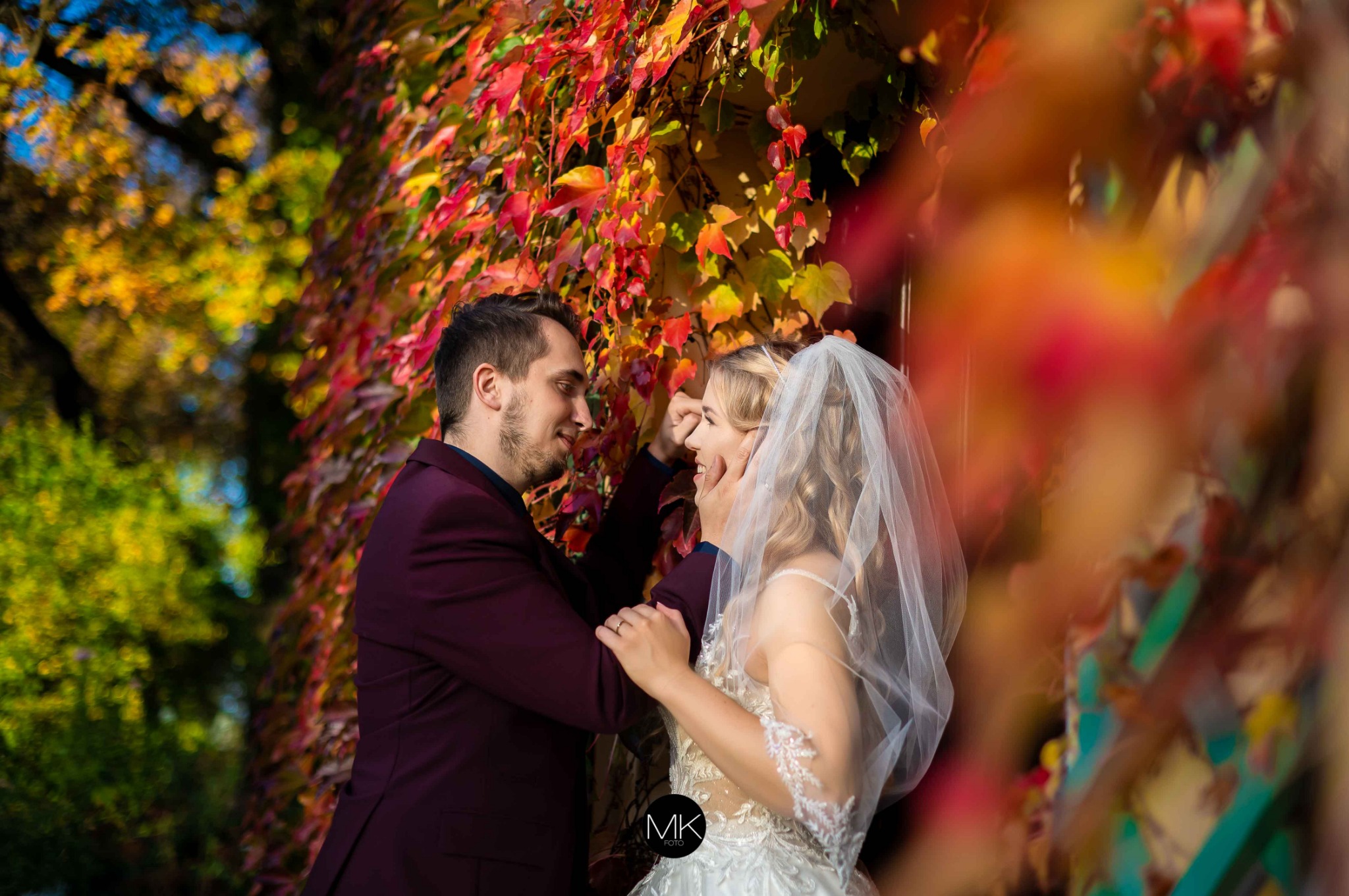 Para młoda w objęciach na tle ściany z kolorowych liści winobluszczu, panna młoda w sukni z koronką i welonem, pan młody w bordowym garniturze, delikatny uśmiech i wzajemne spojrzenie.