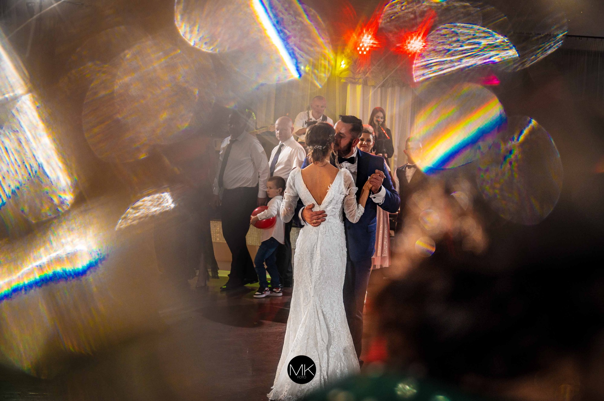 Para młoda w tańcu podczas wesela, panna młoda w koronkowej sukni z odkrytymi plecami, pan młody w granatowym garniturze, w tle zespół muzyczny i goście, efektowne rozmycie światła.