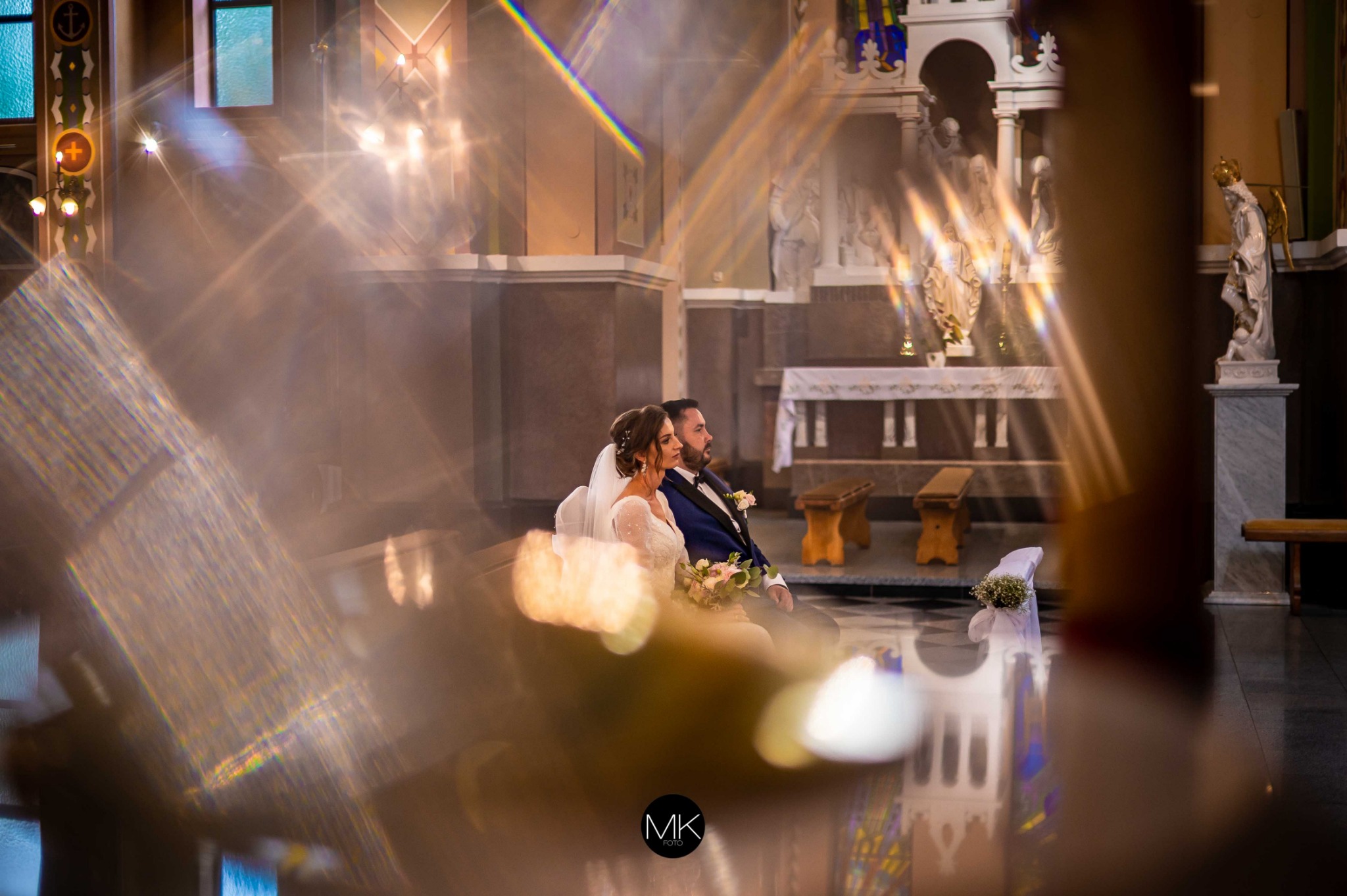Para młoda siedząca w kościele, z efektem załamania światła tworzącym pryzmatyczne refleksy na pierwszym planie. W tle widoczny ołtarz i figury świętych.