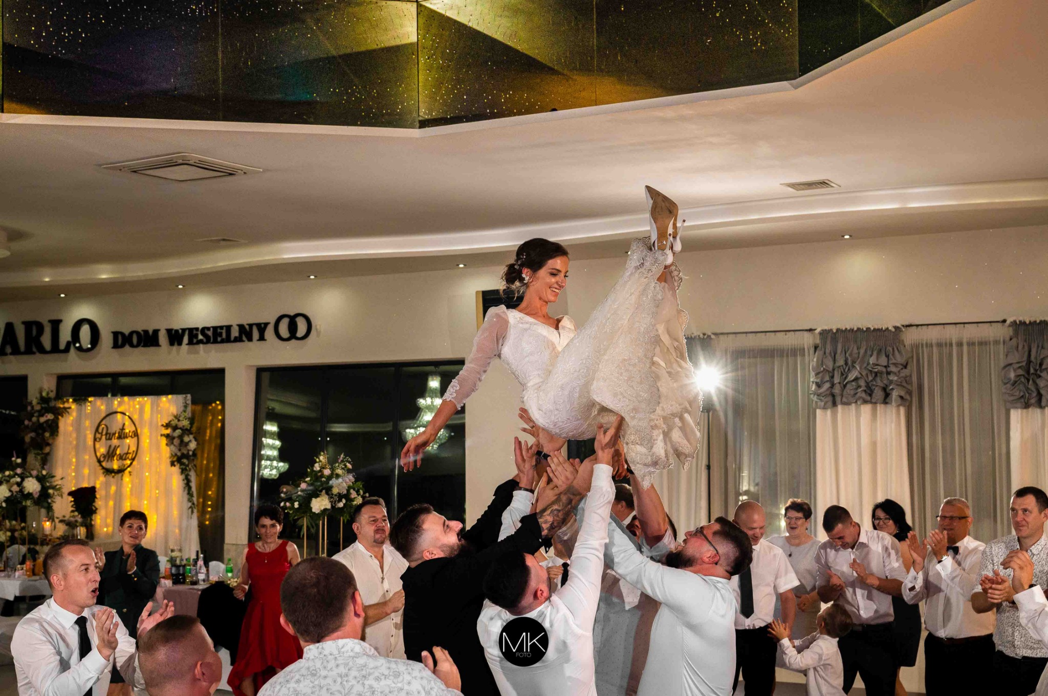 Uśmiechnięta panna młoda w białej sukni, podrzucana do góry przez grupę mężczyzn na sali weselnej z napisem 'Carlo Dom Weselny' w tle.