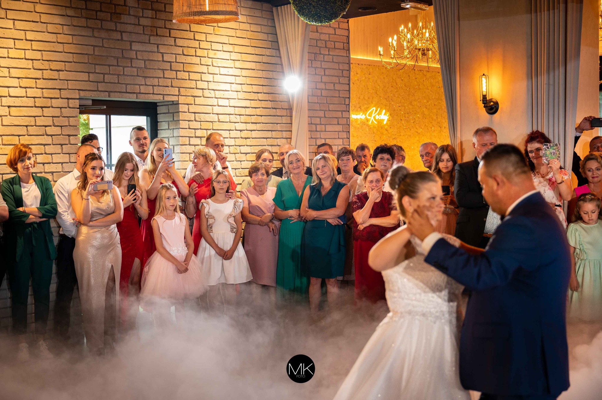 Para młoda podczas oczepin, z tortem na twarzy, otoczona gośćmi weselnymi i sztuczną mgłą na sali z ceglaną ścianą.