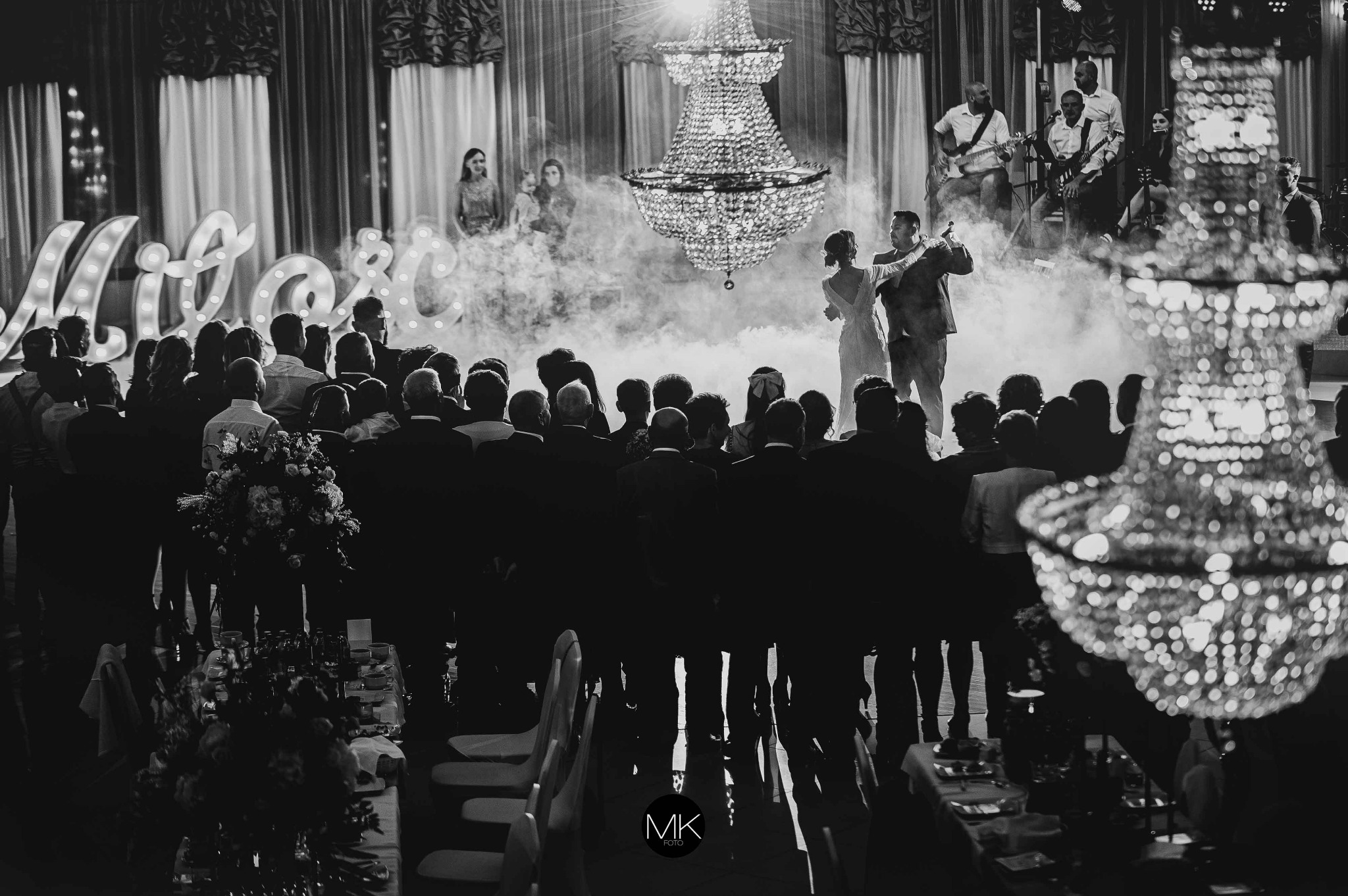 Czarno-białe zdjęcie pary tańczącej w dymie na tle zespołu muzycznego, widzianej zza pleców gości zgromadzonych na sali z kryształowymi żyrandolami i podświetlanym napisem.