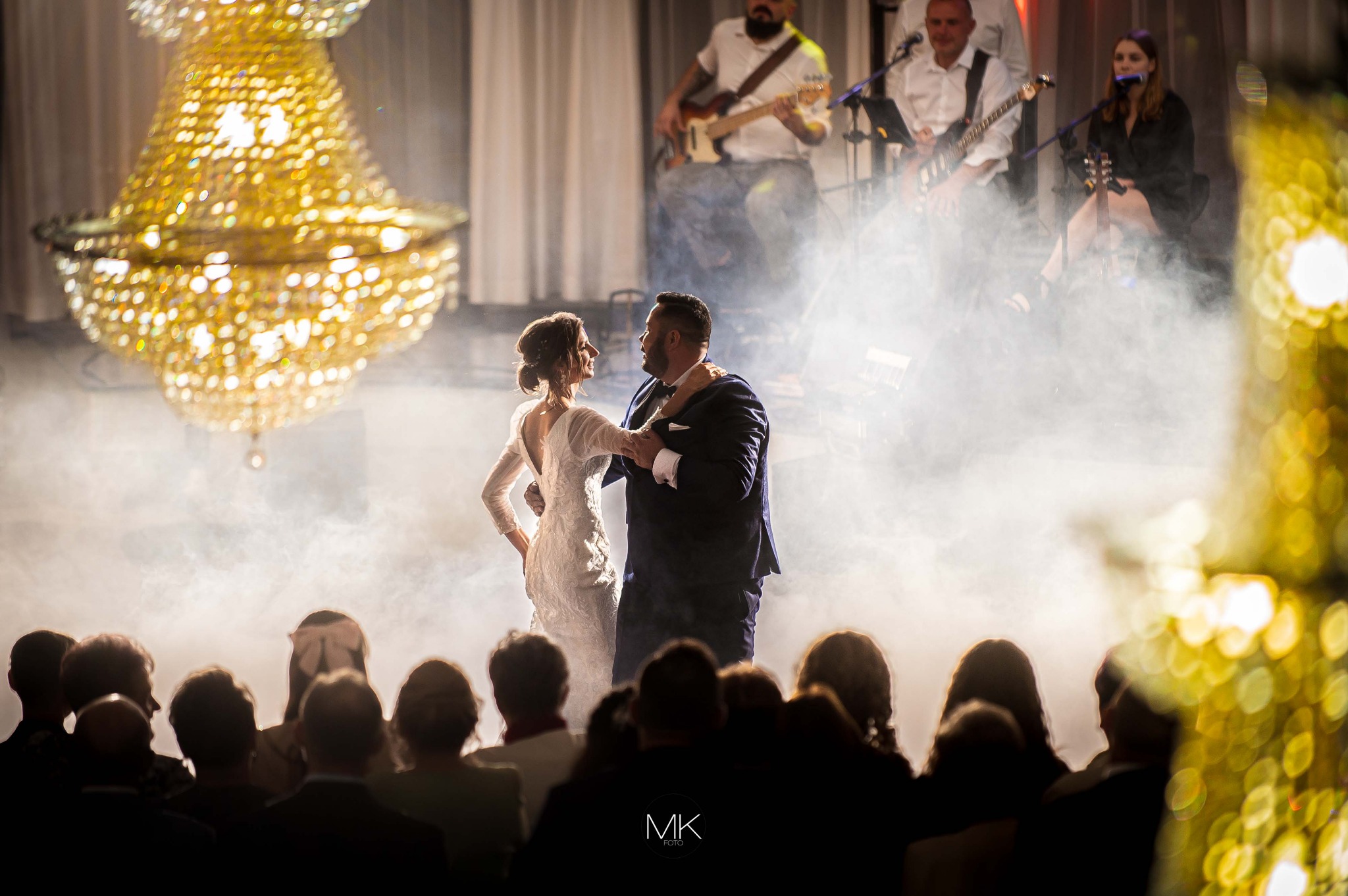 Para młoda tańczy na parkiecie w oparach dymu, za nimi zespół muzyczny i publiczność, nad nimi bogaty żyrandol.